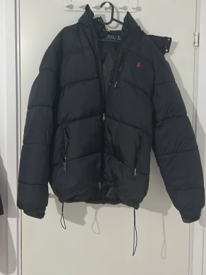Svart pufferjacka från Polo Ralph Lauren - Snygg svart pufferjacka från Polo Ralph Lauren med röd logga på bröstet. Jackan har dragkedja framtill, två sidofickor med dragkedja och justerbar dragsko nedtill. Perfekt för kalla dagar och riktigt stilren look. Storlek S men för lång för mig (är 1.70) använd 3-4 gånger så fortfarande i nyskick