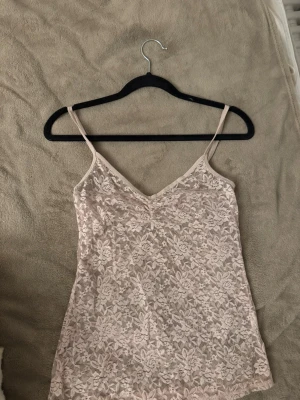 Ljusrosa spetslinne - Säljer ett ljusrosa spetslinne med tunna axelband och v-ringning. Toppen är täckt av vacker spets med blommigt mönster och har en feminin och romantisk look. Perfekt att styla lager på lager eller bära som den är.