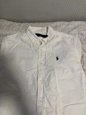 Vit skjorta från Ralph Lauren - Klassisk vit skjorta från Ralph Lauren med button-down krage och blå broderad logga på bröstet. Skjortan har långa ärmar och knappar framtill. Perfekt till jeans eller chinos för en clean och stilren look.