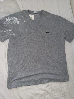 Grå t-shirt från Lacoste - Snygg grå t-shirt från Lacoste med klassisk rund hals och den ikoniska krokodilloggan broderad på bröstet. Tillverkad i mjuk bomull som känns skön mot huden. Perfekt basplagg för en clean och stilren look.