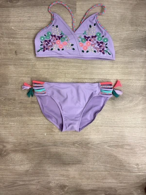 Lila bikini med broderi och tofsar - Superfin lila bikini med färgglada broderade blommor på toppen och flätade axelband. Bikinitrosan har dekorativa band i lila, turkos och rosa samt tofsar på sidorna. Perfekt för sommaren och strandhäng!
