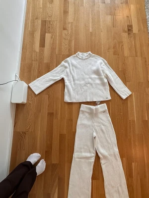 Vitt ribbat loungewear-set - Mysigt loungewear-set i vitt med ribbad struktur. Setet består av en långärmad topp med rund hals och ett par vida byxor med hög midja. Perfekt för chill dagar hemma. Materialet ser mjukt och stretchigt ut.