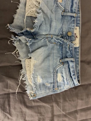 Ljusa jeansshorts med spets Hollister - Ljusa jeansshorts med slitna detaljer, fransar och vit spets i sidorna. Klassisk femficksmodell med knappgylf och råa kanter. Broderade detaljer på bakfickorna och tydlig Hollister-lapp bak. Perfekta för en chill sommarstil.