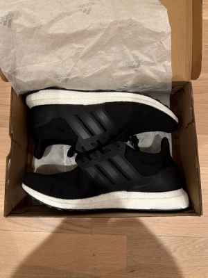 Svarta Adidas Ultra Boost sneakers - Säljer ett par svarta Adidas Ultra Boost sneakers med klassiska tre ränder på sidan. Skorna har en vit, tjock och bekväm sula i Boost-material och ovandel i mesh för bra andningsförmåga. Perfekta för dig som gillar stilrena och sportiga sneakers.