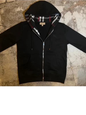 Burberry kofta  - Snygg svart hoodie från Burberry med dragkedja framtill och klassiskt rutigt foder i huvan. Hoodien har två fickor fram och justerbara snören i huvan. Perfekt för dig som vill ha en stilren och exklusiv look med en touch av Burberrys ikoniska mönster. Grejen vid luvan är lite lös men går att fixa 