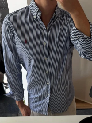 Blårandig skjorta med knappkrage - Snygg skjorta med smala blå och vita ränder, klassisk button-down krage och långa ärmar. Skjortan har broderad logga i rött på bröstet och vita knappar framtill. Perfekt till jeans eller chinos för en clean och stilren look.