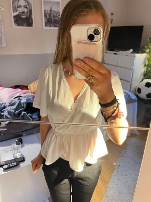 Vit omlottblus med volang - Snygg vit omlottblus med korta ärmar och volang nertill. Blusen har v-ringning med spetsdetalj längs kanten och resår i midjan för en feminin siluett. Perfekt att styla med både jeans och kjol för en trendig look. Lappar är avklippta men jag skulle säga att den är i ca storlek 34.