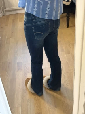 Mörkblå bootcut jeans - Säljer ett par mörkblå bootcut jeans med klassisk femficksdesign och lätt slitna detaljer. Jeansen har normal midja och är tillverkade i ett stretchigt denimtyg som sitter snyggt på benen. Perfekta till en avslappnad look.