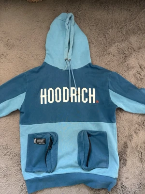 Blå hoodie från Hoodrich med fickor - Säljer en blå hoodie från Hoodrich med stor logga framtill. Hoodien har två stora fickor med dragkedja på magen och justerbar huva med snören. Färgen är en mix av ljus- och mörkblå toner. Perfekt för dig som gillar streetwear och vill sticka ut.