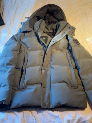 Canada goose vinterjacka - Säljer en ljusgrå pufferjacka med stor huva och quiltad design. Jackan har dragkedja och tryckknappar framtill samt en patch på ärmen. Perfekt för kalla vinterdagar och riktigt skön att bära. Fylld med vaddering för extra värme.