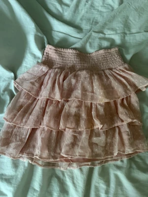 Ljusrosa volangkjol med prickar - Supersöt ljusrosa kortkjol med flera volanger och diskret prickigt mönster. Kjolen har bred resår i midjan och är gjord i ett tunt, luftigt material som passar perfekt till varmare dagar. Perfekt för dig som gillar romantisk och feminin stil.Liten fläck men inget som syns.
