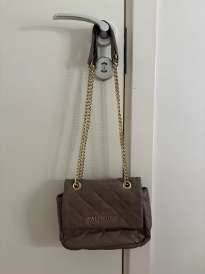 Rosa quiltad handväska från Valentino - Snygg rosa quiltad handväska från Valentino med guldfärgade kedjehandtag och logga framtill. Väskan har en klassisk rektangulär form och är tillverkad i ett mjukt material som ger en lyxig känsla. Perfekt accessoar för att lyfta din outfit.