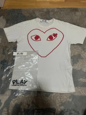 Vit Comme des Garçons Play t-shirt - Vit t-shirt från Comme des Garçons Play med stort rött hjärta och ögon på bröstet. Klassisk rund hals och korta ärmar. Ikonisk design som sticker ut och passar perfekt till streetwear-stilen.