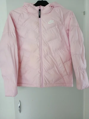 Ljusrosa pufferjacka från Nike - Supersnygg ljusrosa pufferjacka från Nike med huva och dragkedja framtill. Jackan har en klassisk quiltad design och Nike-logga på bröstet. Perfekt för kalla vinterdagar och riktigt skön att bära. Materialet är lätt och håller värmen bra.