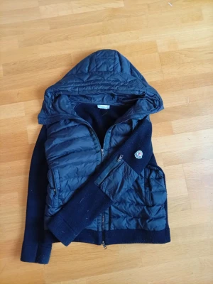 Svart cardigan från Moncler - Snygg svart pufferjacka från Moncler med stickade ärmar och huva. Jackan har dragkedja framtill, två fickor och Moncler-logga på ena ärmen. Perfekt för kalla vinterdagar och har en modern, sportig look.