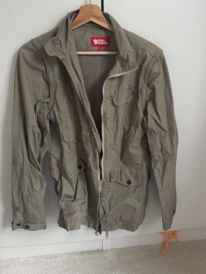 Beige vindjacka från Fjällräven - Stilren beige vindjacka från Fjällräven med dragkedja, flera fickor framtill och meshfoder på insidan.