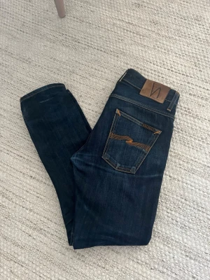  Nudie Jeans DRY SELVEDGE - Säljer ett par mörkblå jeans från Nudie Jeans med klassisk femficksmodell och kontrastsömmar i orange. Modellen är grim tim och de är i storlek W29/L30. De är i materialet dry selvedge och har en fin wash. Hör av dig vid frågor elr liknande!