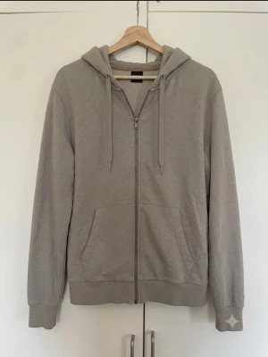 Ljusgrå zip hoodie med huva - En stilren ljusgrå hoodie med dragkedja framtill och två fickor. Tröjan har huva med dragsko och ribbade muddar vid ärmar och nederkant. Perfekt för en avslappnad look och enkel att matcha med andra plagg.
