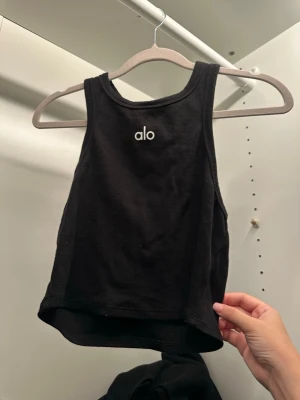  linnetopp från Alo - Snygg svart ribbad linnetopp från Alo med hög halsringning och ärmlös design. Perfekt för träning eller som ett stilrent basplagg. Använd 1 gång. 