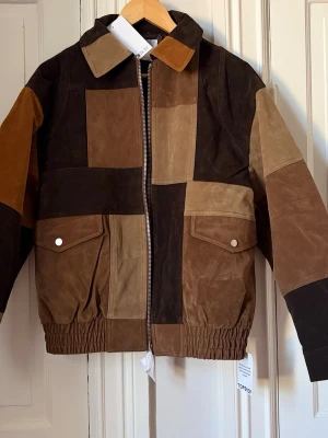 Patchwork skinnjacka i bruna toner - Unik skinnjacka (mocka imitation)  i patchwork-design med olika nyanser av brunt och beige. Jackan har två stora fickor med lock och tryckknappar, dragkedja framtill och elastisk midja. Perfekt för dig som vill sticka ut med en retroinspirerad look. Strl XS men oversize, passar S och även M