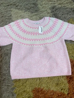 Ljusrosa stickad tröja Kappahl - Supersöt ljusrosa stickad tröja från Kappahl med rund hals och mönster i vitt och rosa runt oket. Tröjan har korta ärmar och ribbade muddar vid ärmslut och nederkant. Perfekt för kalla dagar och riktigt mysig att ha på sig.