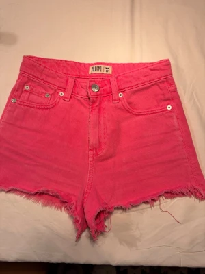 Högmidjade rosa jeansshorts - Säljer ett par  högmidjade jeansshorts i stark rosa färg. Shortsen har råa, fransiga kanter och klassisk femficksdesign.  Storlek 32