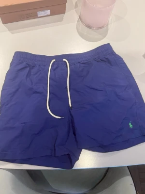 Blå badbyxor från Polo Ralph Lauren - Snygga blå badbyxor från Polo Ralph Lauren med vit dragsko i midjan och klassisk grön logga broderad framtill. Modellen har kortare ben och en bakficka med knapp. Perfekta för stranden eller poolen.