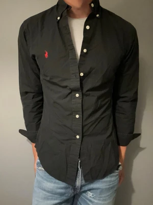 Svart skjorta från Ralph Lauren - Snygg svart skjorta från Ralph Lauren med klassisk button-down krage och vita knappar. Skjortan har lång ärm och den ikoniska röda logotypen broderad på bröstet. Tillverkad i ett tunt och bekvämt bomullstyg som passar perfekt till jeans.