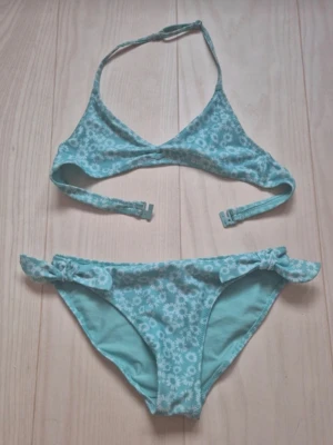 Turkos blommig bikini från H&M - Söt turkos bikini från H&M med vita blommor och volangdetaljer på bikinitrosan. Perfekt för stranddagar och poolhäng. Bikinin har klassisk passform och är tillverkad i mjukt syntetmaterial.