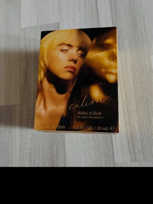 Eilish Billie - Eilish Billie Eilish Eau de Parfum. Förpackningen är guld- och svartfärgad med ett porträtt av Billie Eilish på framsidan. Parfymen är en ny doft och kommer i en elegant, rektangulär kartong.