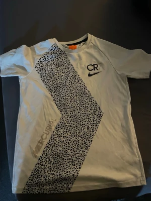 Vit CR7 Nike t-shirt med mönster - Snygg vit t-shirt från Nike CR7-kollektionen med svart prickigt mönster i diagonal design över framsidan. T-shirten har korta ärmar, rund halsringning och är tillverkad i Dri-FIT polyester för en sportig känsla.
