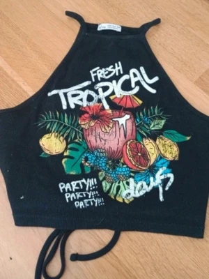 Svart croppad linnetopp med tryck - Säljer en supersöt svart sommartopp med öppen rygg och somrigt tryck från Bershka. 💞 Helt ny och då aldrig använd, men med bortklippta lappar. Skriv om du har någon fråga 🤗