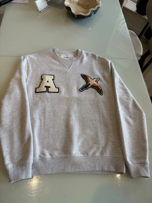 Grå sweatshirt med applikationer - Snygg grå sweatshirt med rund hals och ribbade muddar. På bröstet finns en stor vit bokstav 'A' och en broderad fågel i färg. Perfekt till jeans eller joggers för en avslappnad look.