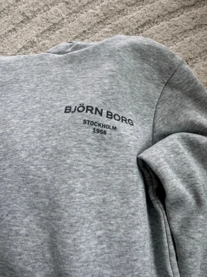 Grå hoodie från Björn Borg - Snygg grå hoodie från Björn Borg med svart tryck på bröstet där det står 'BJÖRN BORG STOCKHOLM 1956'. Mjuk insida och klassisk huva med dragsko. Perfekt för en chill och sportig stil.