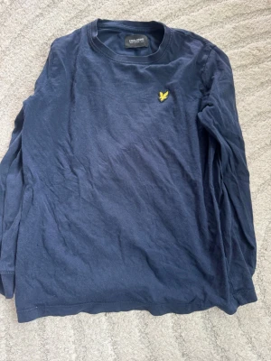 Mörkblå långärmad tröja Lyle & Scott - Mörkblå långärmad tröja från Lyle & Scott Junior med rund hals och den klassiska gula loggan broderad på bröstet. Tröjan är i mjukt bomullsmaterial och passar perfekt till en avslappnad stil. Storlek: 8/9 år