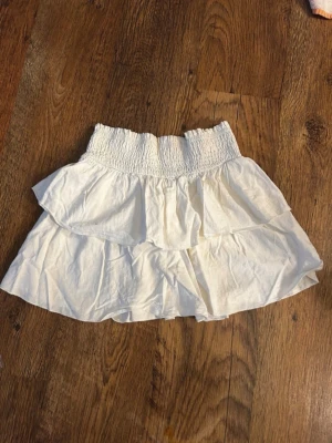 Vit volangkjol med smock - Söt vit kortkjol med bred smock i midjan och dubbla volanger. Kjolen är gjord i ett lätt bomullstyg som känns luftigt och perfekt för varma dagar. Enkel att matcha med olika toppar för en fräsch look.
