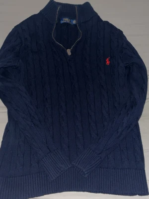 Polo Ralph Lauren Quarter Zip - Mörkblå kabelstickad tröja från Polo Ralph Lauren med halv dragkedja och röd broderad logga på bröstet. Tröjan har ribbade muddar och hög krage, perfekt för lager på lager. Klassisk och tidlös design i mjukt material.