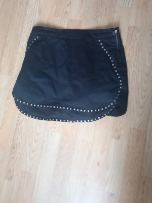 Svart kortkjol med nitar - Snygg svart kortkjol med rundad nederkant och dekorativa silverfärgade nitar längs kanterna. Kjolen har en enkel design med dragkedja i sidan och passar perfekt till en trendig vardagslook.