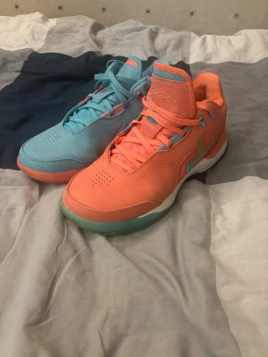 Nike sportskor i blått och orange - Säljer ett par sportskor från Nike där ena skon är ljusblå och den andra är orange. Skorna har snörning, vit sula med turkosa detaljer och en tydlig Swoosh-logga på sidan. Materialet är en mix av läder och textil, perfekt för dig som gillar färgstarka sneakers.