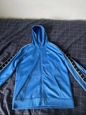 Blå Nike hoodie med dragkedja - Snygg blå hoodie från Nike med hel dragkedja och huva. Ärmarna har svarta detaljer med vita Nike-loggor längs sidorna. Knappt använd så väldigt fint skick. Storlek 158 - 170cm men sitter som XS