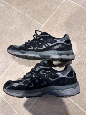 Svarta Asics sneakers med grå detaljer - Snygga svarta Asics sneakers med grå detaljer och reflekterande inslag. Skorna har en sportig design med mesh och syntetmaterial, samt en robust sula för extra komfort. Perfekta för dig som gillar streetwear och sportig stil.