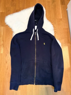 Marinblå hoodie från Polo Ralph Lauren - Snygg marinblå hoodie från Polo Ralph Lauren med dragkedja framtill, vit snörning i huvan och klassisk gul logga på bröstet. Tillverkad i mjuk bomull och har två fickor framtill. Tröjan är i mycket fint skick. Hör av er med frågor & funderingar 🙏