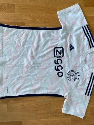 Vit Ajax fotbollströja Adidas - Säljer en vit Ajax fotbollströja från Adidas med marinblå detaljer och tre ränder på axlarna. Tröjan har Ajax-logga och Ziggo-tryck på bröstet samt ett mönster i ljusblått och rosa. Perfekt för träning eller match!