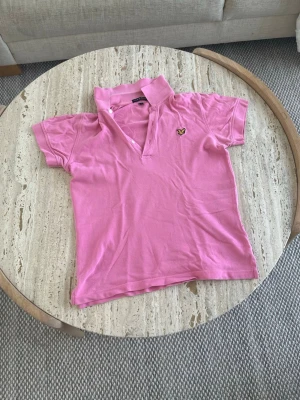 Rosa piké t-shirt med krage - Snygg rosa piké t-shirt med klassisk krage och korta ärmar. På bröstet finns en liten broderad logga i gult och svart. Perfekt för en avslappnad men ändå stilren look.