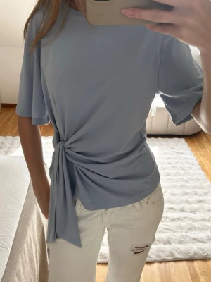 Ljusblå topp med knyt - Superfin ljusblå topp med korta ärmar och snygg knytning i sidan. T-shirten har en avslappnad passform och är gjord i ett mjukt, stretchigt material som känns skönt mot huden. Perfekt att styla med jeans eller kjol för en trendig look.