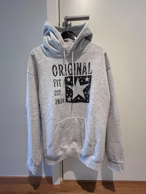 Grå hoodie Gildan  - Grå hoodie med svart tryck framtill där det står 'ORIGINAL FIT EST. 2024' och en stor stjärna med mindre stjärnor runt om. Storlek M men är mer som Small. Perfekt för en avslappnad stil.
