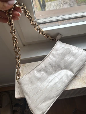 Vit croco-mönstrad handväska med kedja - Snygg vit handväska i croco-mönstrat konstläder med en chunky guldfärgad kedja som axelrem. Väskan har dragkedja upptill och svart innerfoder. Perfekt accessoar för att lyfta din outfit.