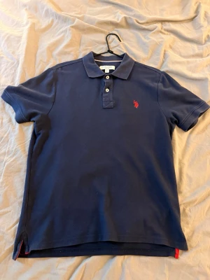 Marinblå pikétröja från U.S. Polo Assn - Klassisk marinblå pikétröja från U.S. Polo Assn med krage, två knappar och röd broderad logga på bröstet. Tröjan har korta ärmar och små röda detaljer vid slitsarna nedtill. Perfekt till jeans eller shorts. Denna har jag använt i ett halvår men jag har varit lite försiktig med den, används inte längre. Nypris 899kr