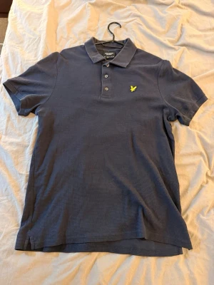 Blå pikétröja från Lyle & Scott - Klassisk blå pikétröja från Lyle & Scott med krage, tre knappar och den ikoniska gula logon broderad på bröstet. Tröjan har korta ärmar och är tillverkad i mjuk bomull för en bekväm känsla.