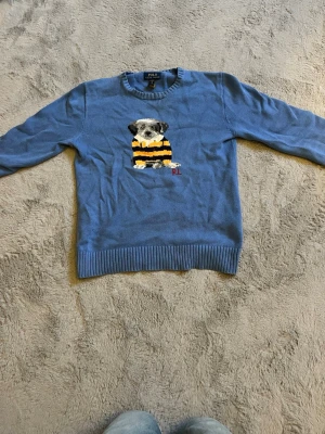 Blå stickad tröja med hundmotiv - Blå stickad tröja från Polo Ralph Lauren med ett broderat hundmotiv på bröstet. Hunden har en gul och svart randig tröja och RL-bokstäver broderade under. Tröjan har långa ärmar och ribbade muddar vid ärmslut och nederkant.